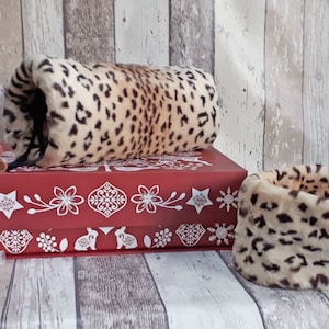 May include: Two leopard print faux fur neck warmers. One is rolled into a tube shape and the other is laid flat. Both are on a red box with white snowflake and floral designs.