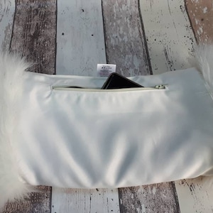May include: White faux fur pillow with a zippered pocket on the back. A black smartphone is inside the pocket.