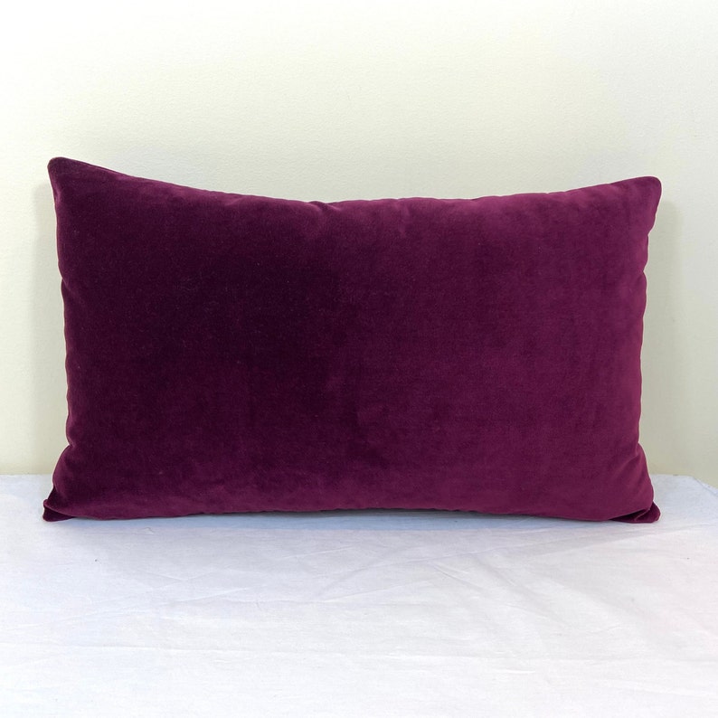 May include: A deep purple velvet pillow with a rectangular shape. The pillow is lying flat on a white surface.