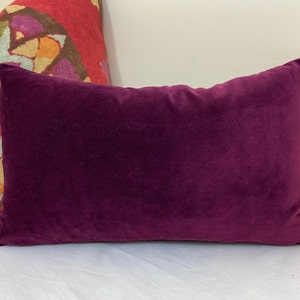 May include: A deep purple velvet pillow with a rectangular shape. The pillow is lying on a white surface.