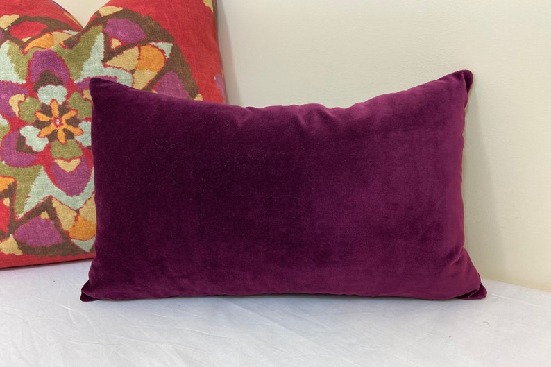 May include: A deep purple velvet pillow with a rectangular shape. The pillow is lying on a white surface.