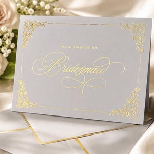 以下が含まれることがあります： 「WILL YOU BE MY Bridesmaid」と金箔で記されたライトグレーの二つ折りカード。ブロック体と筆記体を組み合わせた文字が特徴で、四隅には金箔の繊細な花柄の装飾と枠線が施されています。同色のグレーの封筒が添えられています。