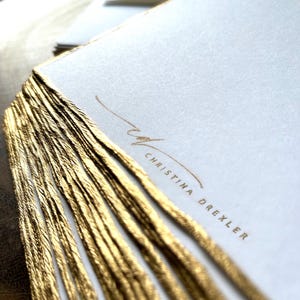 May include: A stack of white stationery cards with gold foil edges. The cards have the name "Christina Drexler" printed in gold lettering.