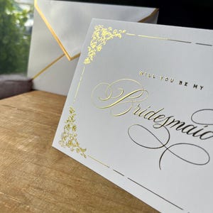 以下が含まれることがあります： 「WILL YOU BE MY Bridesmaid」という文字がエレガントな筆記体とブロック体で記された、ゴールドの箔押しが施された白いカード。カードにはゴールドの装飾的なコーナーデザインが施されており、ゴールドの縁取りがされたお揃いの白い封筒が添えられています。