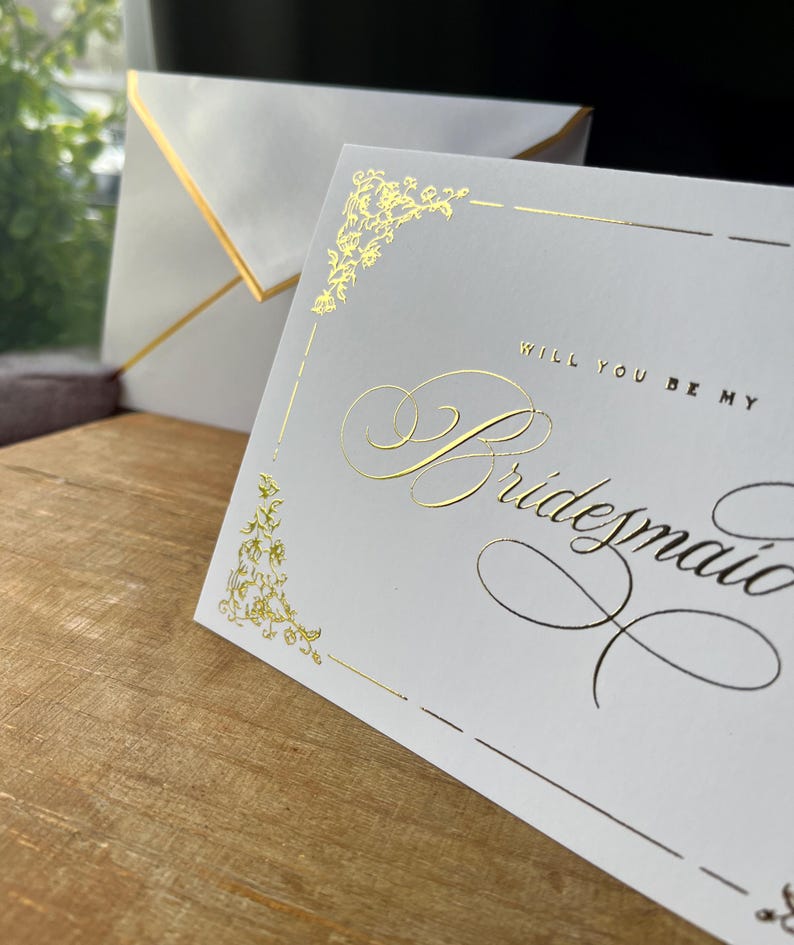 以下が含まれることがあります： 「WILL YOU BE MY Bridesmaid」という文字がエレガントな筆記体とブロック体で記された、ゴールドの箔押しが施された白いカード。カードにはゴールドの装飾的なコーナーデザインが施されており、ゴールドの縁取りがされたお揃いの白い封筒が添えられています。