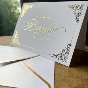 以下が含まれることがあります： Will You Be My Bridesmaidと筆記体で金箔押しされたライトグレーの二つ折りカード。カードの四隅には金箔のフローラルな装飾が施されており、金箔の縁取りがされた同色のライトグレーの封筒が添えられています。