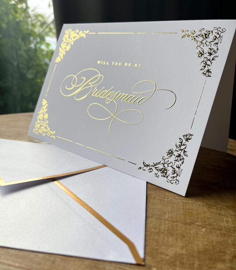 以下が含まれることがあります： Will You Be My Bridesmaidと筆記体で金箔押しされたライトグレーの二つ折りカード。カードの四隅には金箔のフローラルな装飾が施されており、金箔の縁取りがされた同色のライトグレーの封筒が添えられています。