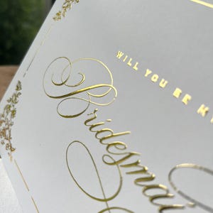 以下が含まれることがあります： 「Will you be my bridesmaid」という文字がエレガントな筆記体とブロック体で金箔押しされた白いカード。カードの四隅には金箔のフローラルな装飾が施され、細い金の縁取り線で囲まれています。