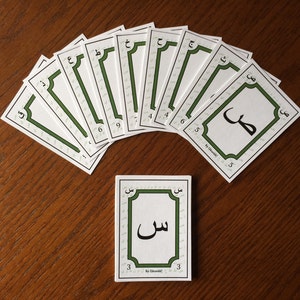 Peut inclure: Un jeu de 10 cartes blanches avec des bordures vertes et des lettres arabes. Les cartes sont disposées en éventail sur une surface en bois. Les lettres sont écrites à l'encre noire. Les cartes sont numérotées de 3 à 9.