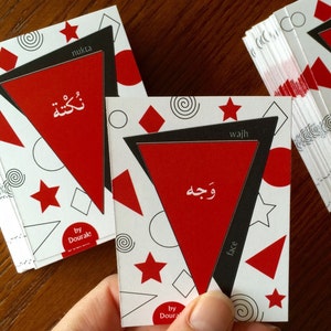 Könnte beinhalten: Zwei rote und schwarze Karten mit arabischem Text und englischen Übersetzungen. Die Karten sind Teil eines Sets namens "Dourak!" und zeigen geometrische Formen in Schwarz und Weiß.