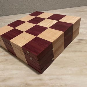 May include: A wooden chessboard with a checkerboard pattern of light wood and dark purple wood. The board is folded in half, revealing the layers of wood.