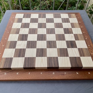 Puede incluir: Un tablero de ajedrez de madera con un patrón de cuadros claros y oscuros. El tablero está enmarcado por un borde de madera más oscura con coordenadas de letras y números. Un juego de mesa clásico de estrategia.