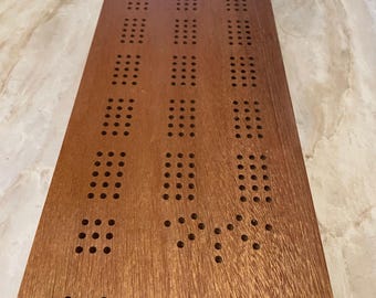 Könnte beinhalten: Ein hölzernes Cribbage-Brett mit natürlicher Oberfläche. Das Brett hat ein traditionelles Design mit Löchern für Stifte und einem Punktezählbereich.