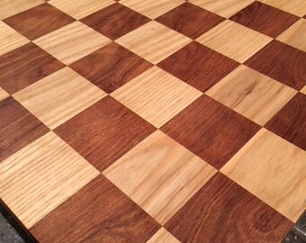 May include: A wooden chessboard with alternating light and dark brown squares. The board is made of two different types of wood, creating a contrasting pattern. The squares are evenly sized and arranged in a classic checkerboard design.