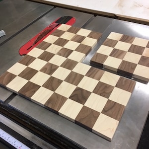 May include: Two wooden chess boards, one larger than the other, are laid out on a table saw. The boards are made of light and dark wood, creating a checkered pattern. The boards are ready to be cut to size.