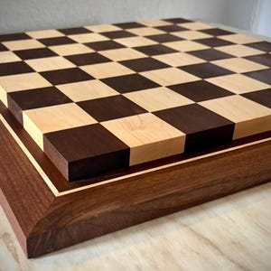 May include: A handcrafted wooden chess board with a checkerboard pattern of light and dark squares. The board is framed with a dark wood border and a lighter wood inlay. The chess board is a classic game for all ages.