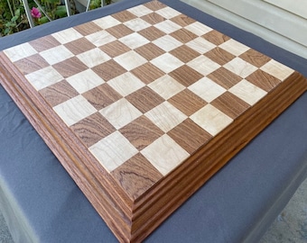 May include: A handcrafted wooden chess board with alternating light and dark squares. The board is framed with a layered, dark brown border. The squares are light and dark wood tones, creating a classic checkerboard pattern.