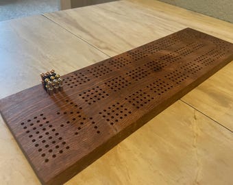 Könnte beinhalten: Ein hölzernes Cribbage-Spielbrett mit zahlreichen Löchern und Metallstiften. Das rechteckige Brett hat eine dunkelbraune Oberfläche. Die Stifte sind gold- und kupferfarben.