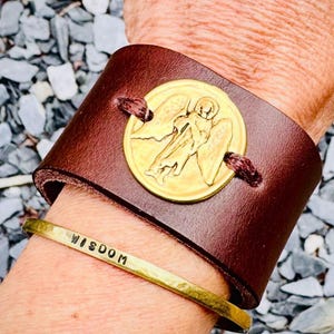 Puede incluir: Una pulsera de cuero marrón con un medallón dorado de ángel y una pulsera dorada con la palabra "WISDOM" estampada. La pulsera se asegura con costuras de color rojo oscuro.