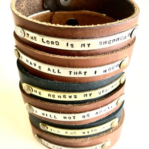 May include: A stack of brown leather bracelets with silver and brass metal bands. Each band has a different inspirational quote engraved on it. The quotes are: "The Lord is my Shepherd", "I have all that I need", "He renews my strength", "I will not be afraid", "You are with me", and "My cup overflows".