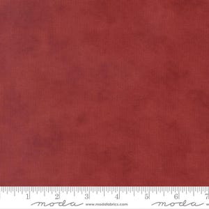 May include: A close-up of a piece of fabric in a solid, muted red-brown color. The fabric has a subtle texture and a ruler is placed along the bottom edge. The fabric is likely intended for quilting or other sewing projects.