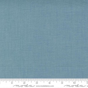 May include: A close-up of a piece of blue fabric with a textured appearance. The fabric has a subtle, woven pattern and a ruler is placed at the bottom. The fabric is a solid color and appears to be cotton or linen.