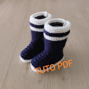 Peut inclure: Une paire de chaussons pour bébé en tricot bleu et blanc avec un revers blanc et deux rayures blanches. Les chaussons sont sur une surface en bois. Le texte "Tuto PDF" est écrit en orange sur l'image.