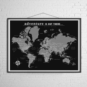 Può includere: Una mappa del mondo in bianco e nero con il testo "adventure is out there..." in alto. La mappa è incorniciata da una cornice nera e appesa a un muro di mattoni bianchi.