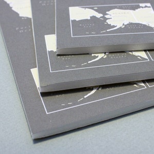 May include: A stack of three gray paper notebooks with white text and maps printed on the covers. The top notebook has a map of Alaska with the text "Gulf of Alaska" and "Seward Peninsula". The middle notebook has a map of Alaska with the text "Gulf of Alaska". The bottom notebook has a map of Alaska with the text "Seward Peninsula".