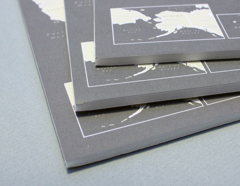 May include: A stack of three gray paper notebooks with white text and maps printed on the covers. The top notebook has a map of Alaska with the text "Gulf of Alaska" and "Seward Peninsula". The middle notebook has a map of Alaska with the text "Gulf of Alaska". The bottom notebook has a map of Alaska with the text "Seward Peninsula".