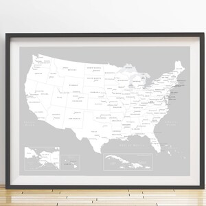 May include: Framed grayscale map of the United States, including Alaska, Hawaii, and the Caribbean. The map is in a black frame and is displayed on a white wall. The map shows state names and major bodies of water.