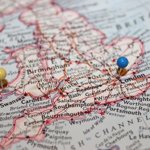 May include: A close-up of a map of the United Kingdom with two push pins. One pin is yellow and is placed on Swansea, Wales. The other pin is blue and is placed on London, England.