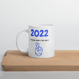Puede incluir: Taza de cerámica blanca con un diseño 2022 azul y el texto "Please don't be ugly" con una mano de dibujos animados azul haciendo un gesto de dedos cruzados.
