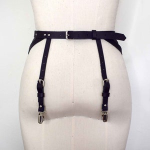 K&ouml;nnte beinhalten: Schwarzer Leder-Strumpfbandg&uuml;rtel mit silbernen Schnallen und Riemen. Der G&uuml;rtel hat ein breites Band und zwei verstellbare Riemen, die an den Beinen befestigt werden.