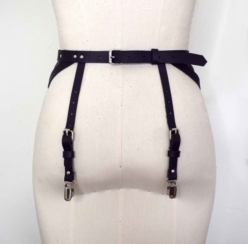 K&ouml;nnte beinhalten: Schwarzer Leder-Strumpfbandg&uuml;rtel mit silbernen Schnallen und Riemen. Der G&uuml;rtel hat ein breites Band und zwei verstellbare Riemen, die an den Beinen befestigt werden.