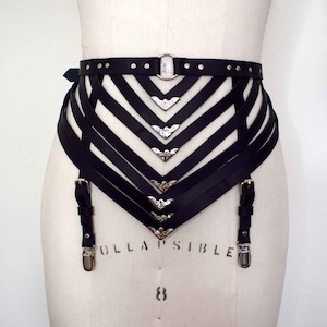 K&ouml;nnte beinhalten: Schwarzer Leder-Strumpfbandg&uuml;rtel mit silbernen Metallakzenten und einer dekorativen Schnalle. Der G&uuml;rtel hat mehrere Riemen und ist f&uuml;r den Tragen um die Taille konzipiert.