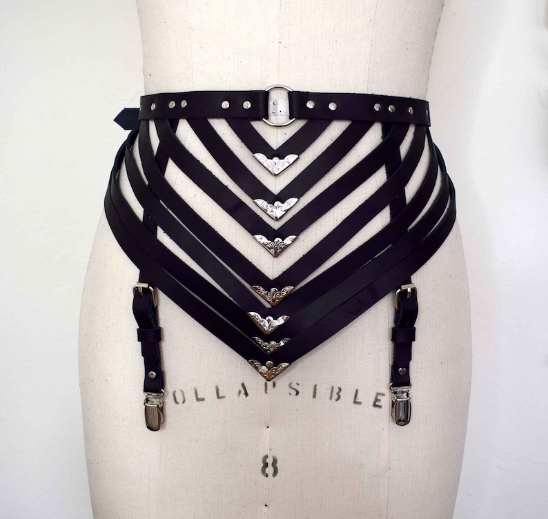 K&ouml;nnte beinhalten: Schwarzer Leder-Strumpfbandg&uuml;rtel mit silbernen Metallakzenten und einer dekorativen Schnalle. Der G&uuml;rtel hat mehrere Riemen und ist f&uuml;r den Tragen um die Taille konzipiert.