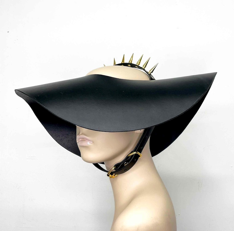 May include: A wide-brimmed black leather hat with a gold spiked headband. The hat has a dramatic, angled brim and a chin strap.