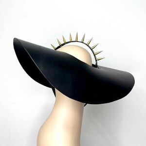 May include: A wide-brimmed black hat with a gold and silver spiked crown. The hat is made of a smooth, black material and has a wide, flat brim. The crown is adorned with a row of gold spikes that are interspersed with silver spikes.