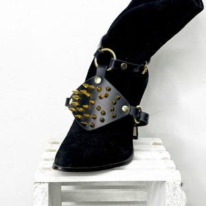 Puede incluir: Bota de ante negra con un accesorio de cuero negro con tachuelas doradas. El accesorio se envuelve alrededor del tobillo y sobre la parte superior del pie, con púas doradas y herrajes dorados. La bota está sobre una superficie de madera blanca.