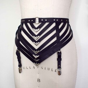 K&ouml;nnte beinhalten: Schwarzer Leder-Strumpfbandg&uuml;rtel mit silbernen Metallakzenten und verstellbaren Tr&auml;gern. Der G&uuml;rtel hat ein dekoratives, geometrisches Design mit mehreren Tr&auml;gern und Schnallen.