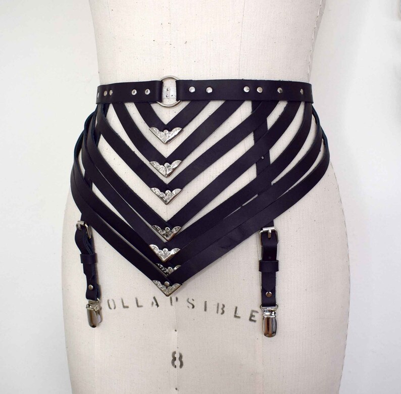 K&ouml;nnte beinhalten: Schwarzer Leder-Strumpfbandg&uuml;rtel mit silbernen Metallakzenten und verstellbaren Tr&auml;gern. Der G&uuml;rtel hat ein dekoratives, geometrisches Design mit mehreren Tr&auml;gern und Schnallen.