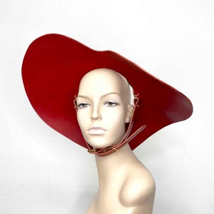May include: A large, red, leather hat with a wide brim and a strap that goes around the neck. The hat is worn on a mannequin.