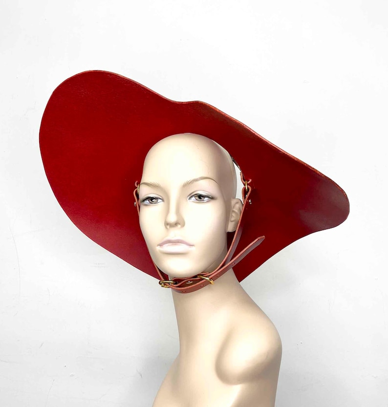 May include: A large, red, leather hat with a wide brim and a strap that goes around the neck. The hat is worn on a mannequin.