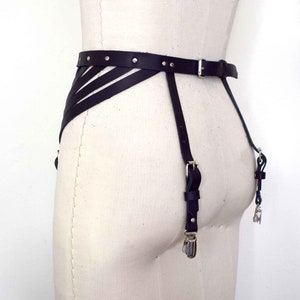 K&ouml;nnte beinhalten: Schwarzer Leder-Strumpfbandg&uuml;rtel mit silberner Hardware und verstellbaren Tr&auml;gern. Der G&uuml;rtel hat ein dekoratives Design mit mehreren Tr&auml;gern, die sich vorne kreuzen.
