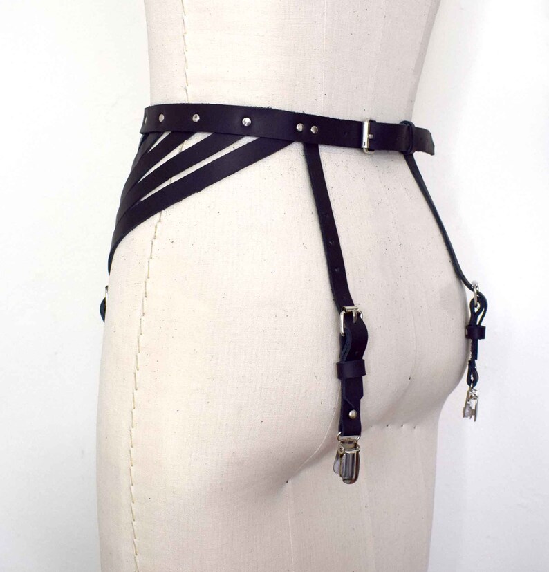 K&ouml;nnte beinhalten: Schwarzer Leder-Strumpfbandg&uuml;rtel mit silberner Hardware und verstellbaren Tr&auml;gern. Der G&uuml;rtel hat ein dekoratives Design mit mehreren Tr&auml;gern, die sich vorne kreuzen.