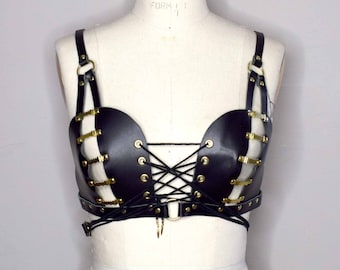 Könnte beinhalten: Ein schwarzer Leder-BH mit goldenen Metall-Akzenten und einem Schnürverschluss vorne. Der BH hat ein Harness-Design mit Trägern, die über die Schultern gehen.