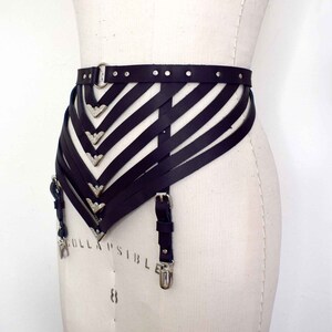 K&ouml;nnte beinhalten: Schwarzer Leder-Strumpfbandg&uuml;rtel mit silbernen Akzenten und verstellbaren Tr&auml;gern. Der G&uuml;rtel hat ein geometrisches Design mit mehreren Tr&auml;gern, die sich kreuzen und eine V-Form bilden.