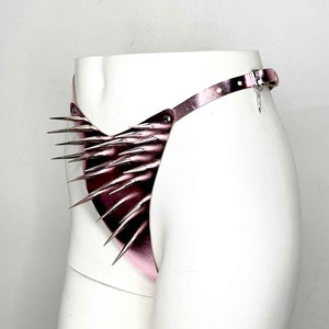 May include: A pink leather body harness with a row of silver spikes. The harness has a strap that goes around the waist and a strap that goes over the shoulder.