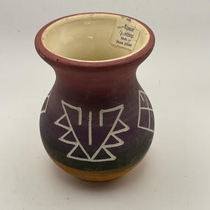 May include: A small, hand-painted ceramic vase with a wide opening and a rounded body. The vase features a design of white geometric shapes on a purple background, with a dark green and orange base. The label reads "Sioux Pottery Made by Sioux Indian."